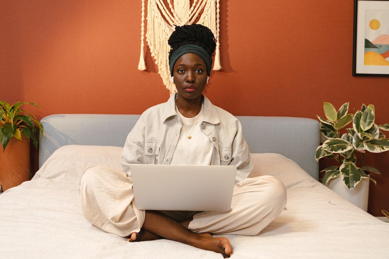 Une femme noir assise dans un lit avec son ordinateur sur les genoux entrain d'ecrire sur la clavier